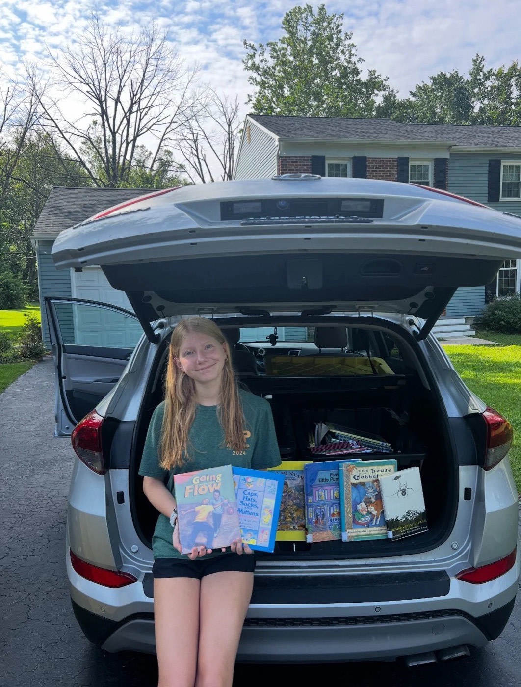 A volunteer holding a stack of donated children's books beside a car trunk full of more books.