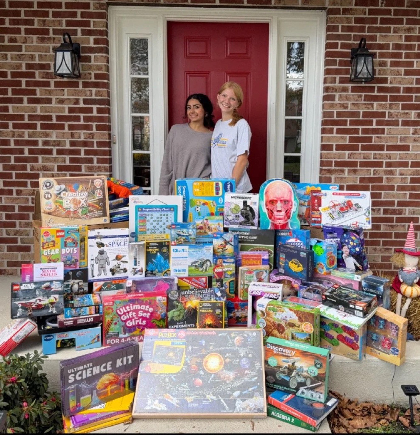 STEM Spark's two founders on a porch beside a large pile of donated science kits and STEM toys from the toy drive.