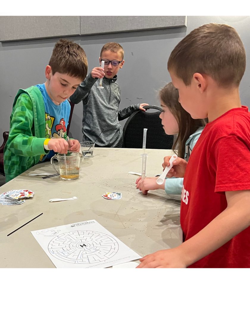 Kids gathered around a table running a hands-on STEM experiment with beakers and syringes at a STEM Spark workshop.